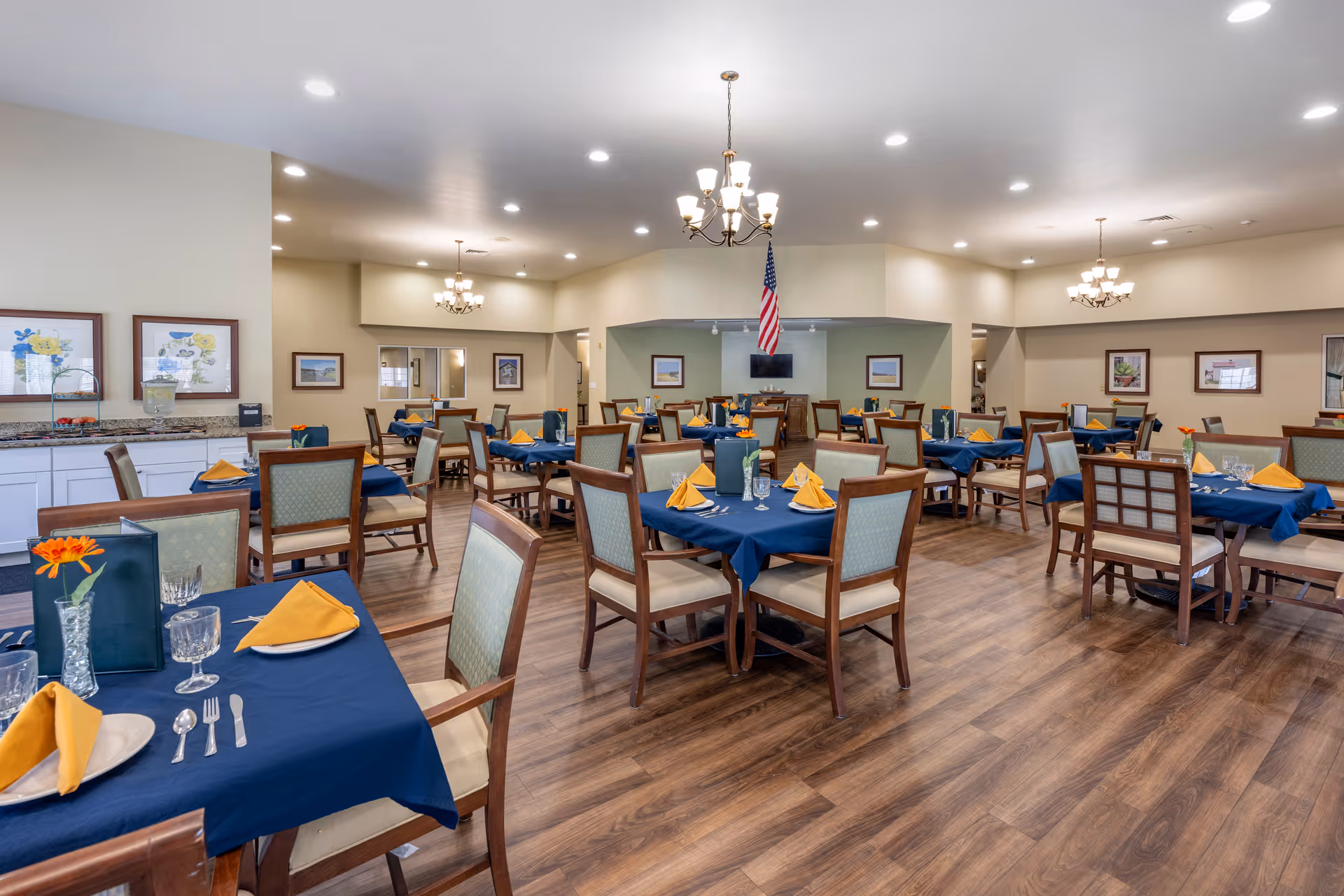 A spacious dining room with multiple tables covered in blue tablecloths and set with yellow napkins, glassware, and silverware. The room features wooden chairs with cushioned seats, wood flooring, beige walls adorned with framed artwork, and several chandeliers hanging from the ceiling. An American flag is displayed at the far end of the room.