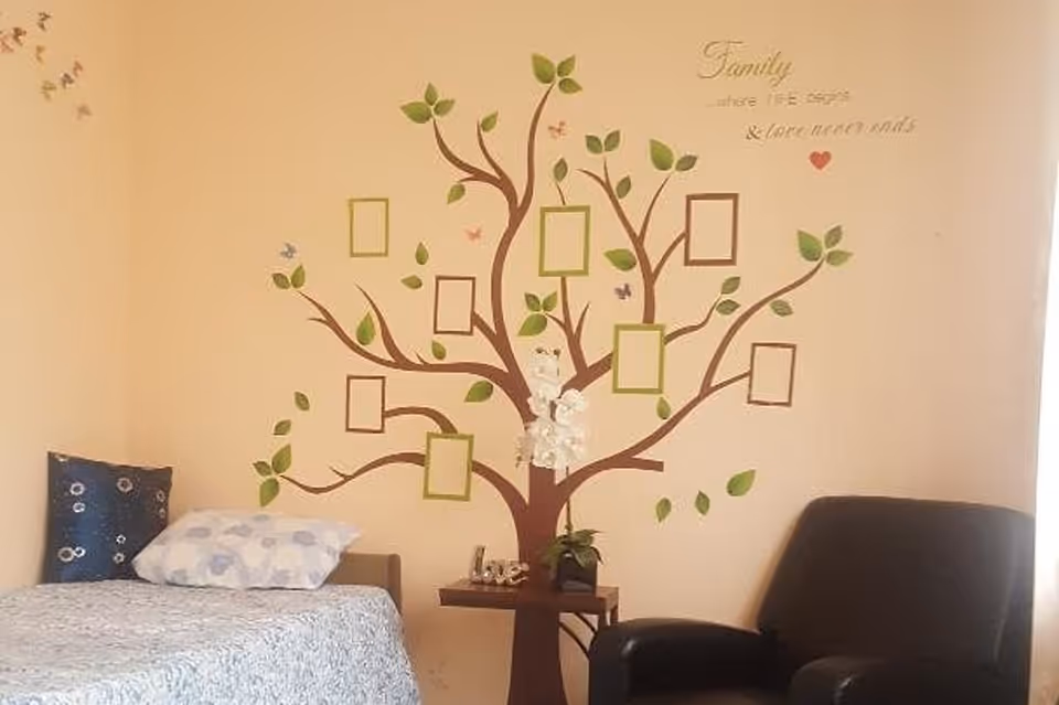 A cozy room with a single bed on the left side, adorned with a blue pillow and a white pillow with blue patterns. On the wall behind the bed is a large tree decal with empty picture frames hanging from its branches and green leaves. There is a small table beneath the tree decal with a white flower arrangement and a small plant. To the right of the table is a dark armchair. Above the tree decal, there is a quote that reads, 'Family where life begins & love never ends.'