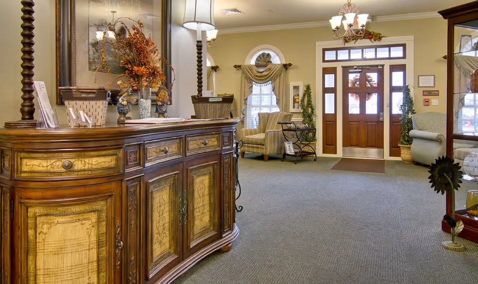 A warmly lit interior lobby area with a decorative wooden sideboard featuring intricate designs and two lamps. In the background, there are comfortable armchairs near windows with draped curtains, a glass display cabinet, and a wooden door with glass panels. The space is carpeted and decorated with plants and autumn-themed floral arrangements.