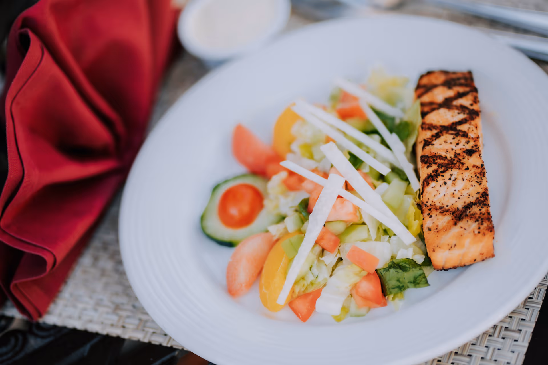 A white plate with a grilled salmon fillet and a fresh salad consisting of lettuce, tomato, cucumber, and thin white vegetable strips, placed on a woven placemat next to a folded red cloth napkin.
