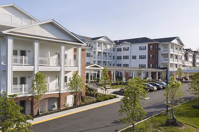 Exterior view of a senior living facility with multiple white buildings featuring balconies, a covered entrance, landscaped greenery, and a parking area with several cars.