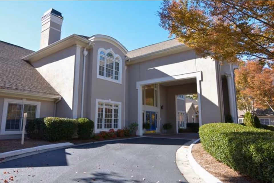 Exterior view of a senior living facility building with beige walls, multiple windows, a covered entrance, and surrounding greenery including bushes and trees with autumn foliage.
