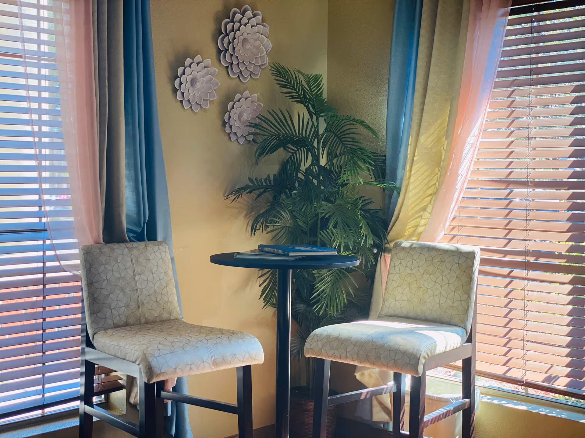 Two upholstered chairs and a small round table beside a potted plant in a sunlit seating area with window blinds and curtains.