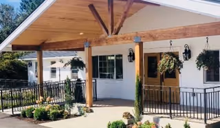 Covered entrance area of a building with wooden beams and a sloped roof, featuring hanging flower baskets, black metal railings, and a pathway surrounded by greenery and flowers.