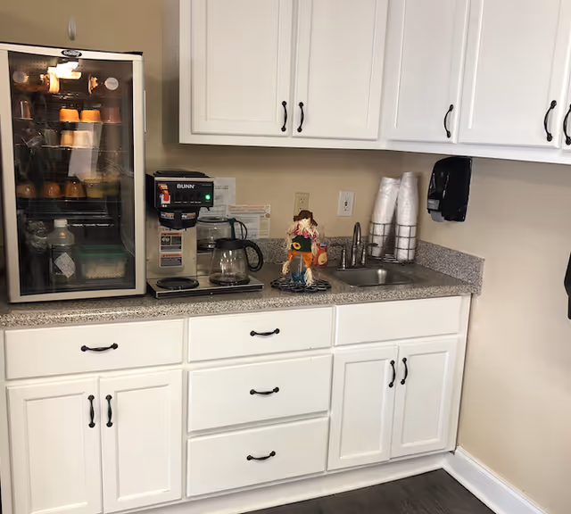 A kitchenette area with white cabinets and drawers, a granite countertop, a small sink, a coffee maker, a mini fridge with various items inside, a stack of disposable cups, a wall-mounted soap dispenser, and a small decorative scarecrow figure on the counter.
