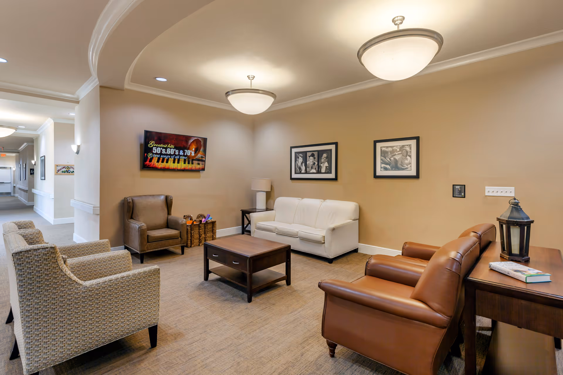 A cozy living room area in a senior living facility with a white sofa, two brown leather armchairs, two patterned armchairs, a brown armchair, a wooden coffee table, and a side table with a lamp and a lantern. The walls are beige with two black and white framed pictures and a flat-screen TV displaying a colorful image. The room has soft overhead lighting and an open hallway visible in the background.