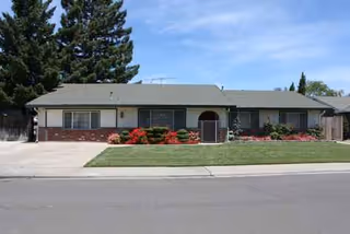 Single-story beige building with gray roof, front lawn, red shrubs, and driveway.