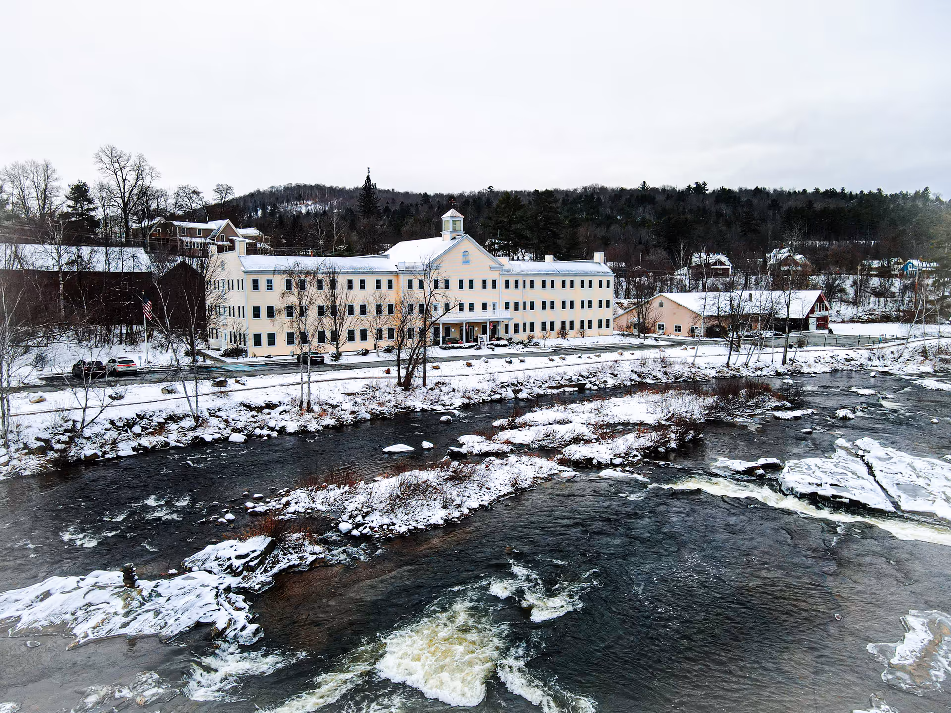 A large, multi-story beige building with many windows situated beside a flowing river with snow-covered banks. The surrounding area is snowy with leafless trees and hills in the background under a cloudy sky.