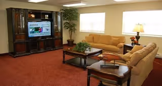 A cozy living room with two beige sofas, a wooden coffee table with a plant centerpiece, a wooden side table with a lamp and decorative items, a large dark wood entertainment center with a television displaying an agenda, and a potted plant in the corner near two windows with blinds.