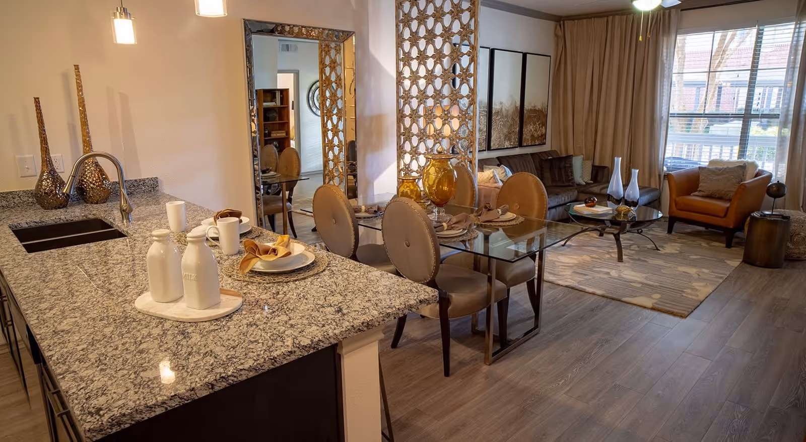 A modern open-concept living space featuring a kitchen island with granite countertop and sink, set with plates, cups, and napkins. Adjacent to the kitchen is a glass dining table with four upholstered chairs. The living room area includes a sofa, armchair, coffee table, and large windows with curtains allowing natural light to fill the room. Decorative elements include vases, a large mirror, and a patterned room divider.