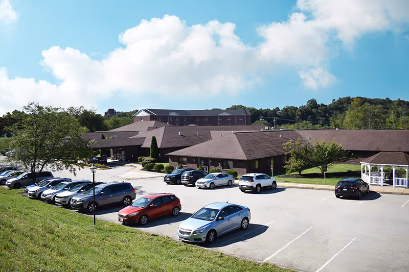 Exterior view of Loyalhanna Care Center showing a large parking lot with several parked cars, a single-story building with a brown roof, green trees, and a blue sky with clouds in the background.