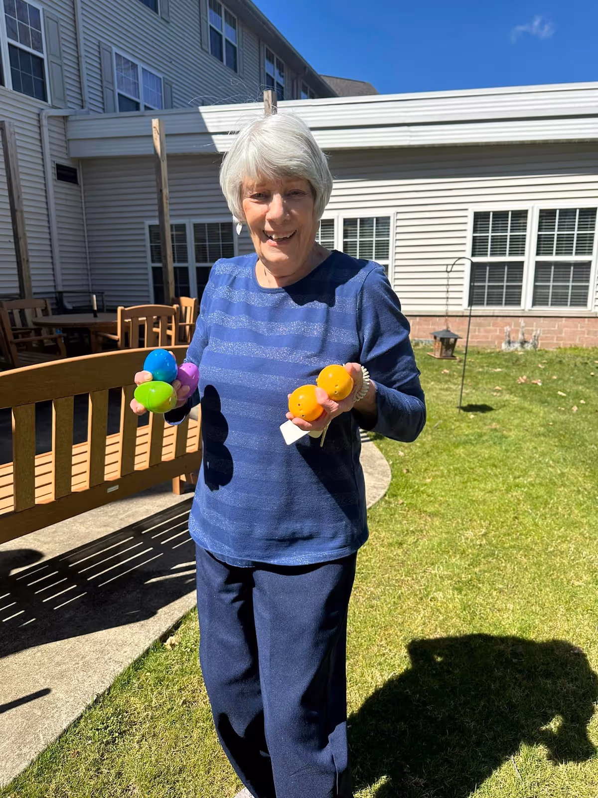 An elderly woman with short white hair wearing a blue striped long-sleeve shirt and dark pants stands outside on a grassy area holding colorful plastic Easter eggs in both hands. Behind her is a building with white siding and multiple windows, along with outdoor wooden benches and tables. The sky is clear and blue.
