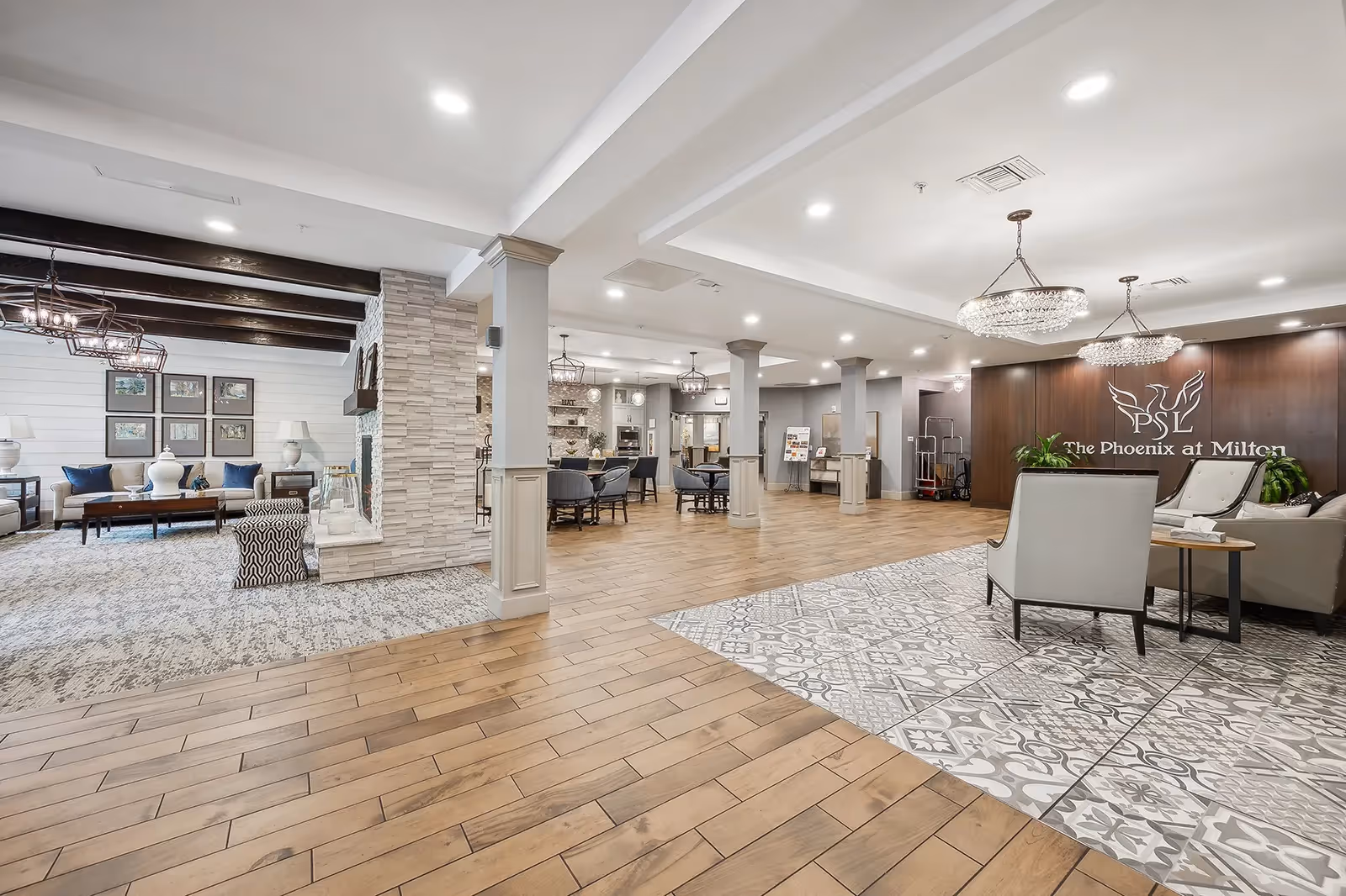Spacious, well-lit senior living lobby with seating areas, chandeliers, wood and patterned tile flooring, and a 'The Phoenix at Milton' sign on a wood-paneled wall.