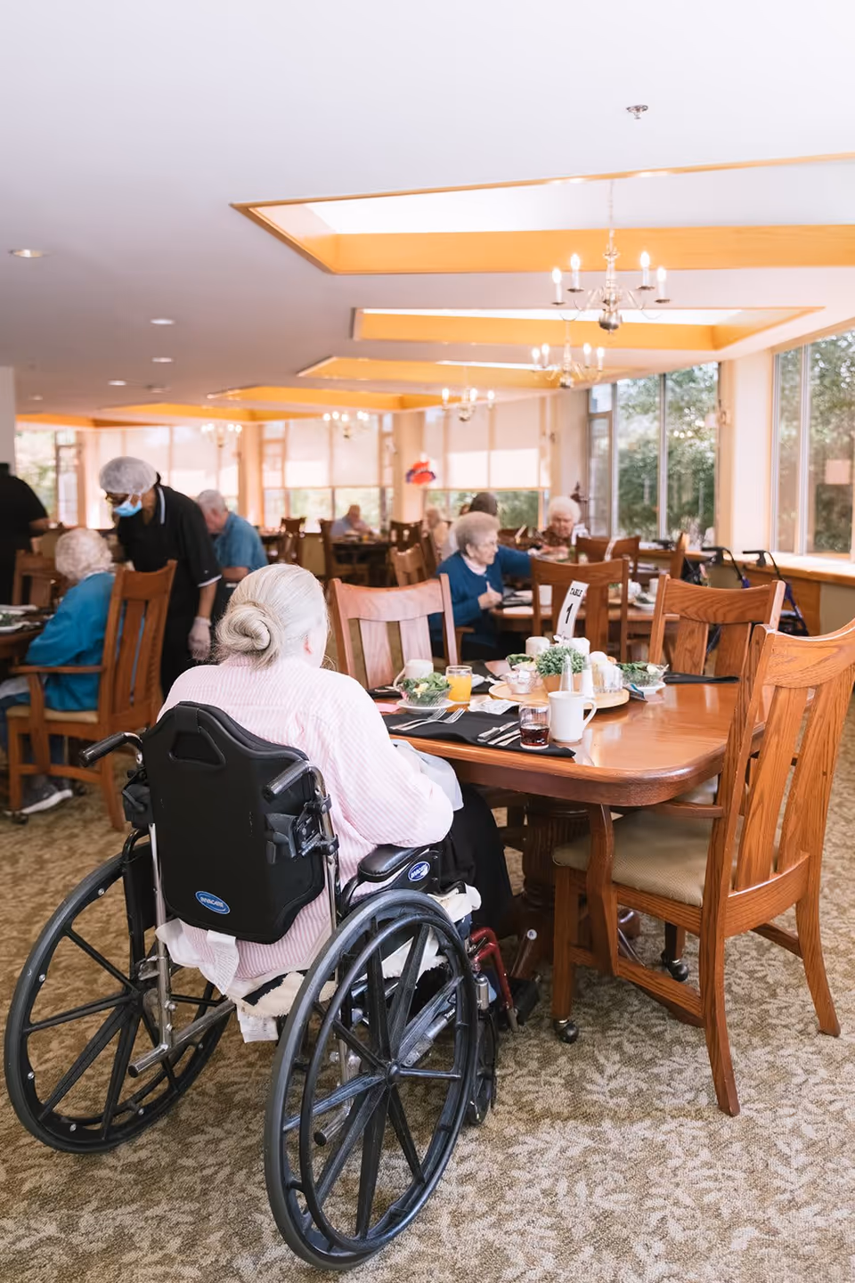 A dining room in a senior living facility with elderly residents seated at wooden tables. One elderly woman in a wheelchair is in the foreground, facing away from the camera. Other residents are seated at tables in the background, and a staff member wearing a hairnet and mask is assisting. The room has large windows letting in natural light and chandeliers hanging from the ceiling.