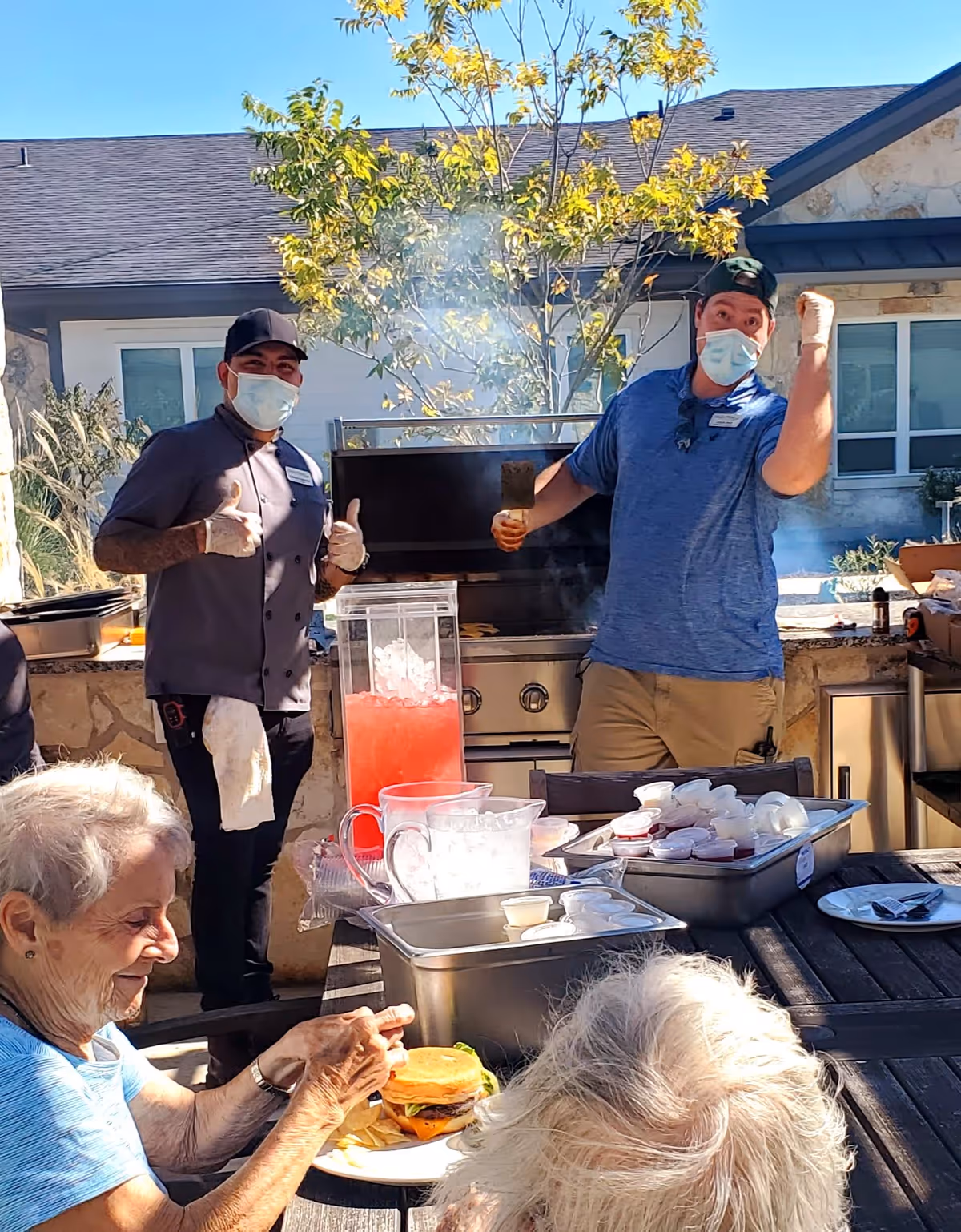 Two men wearing face masks and gloves stand behind an outdoor grill with smoke rising, giving thumbs up and a fist pump. In the foreground, two elderly women sit at a table with food and drinks, including a burger and fries. The setting is an outdoor patio area with a stone wall and a tree with yellow leaves.