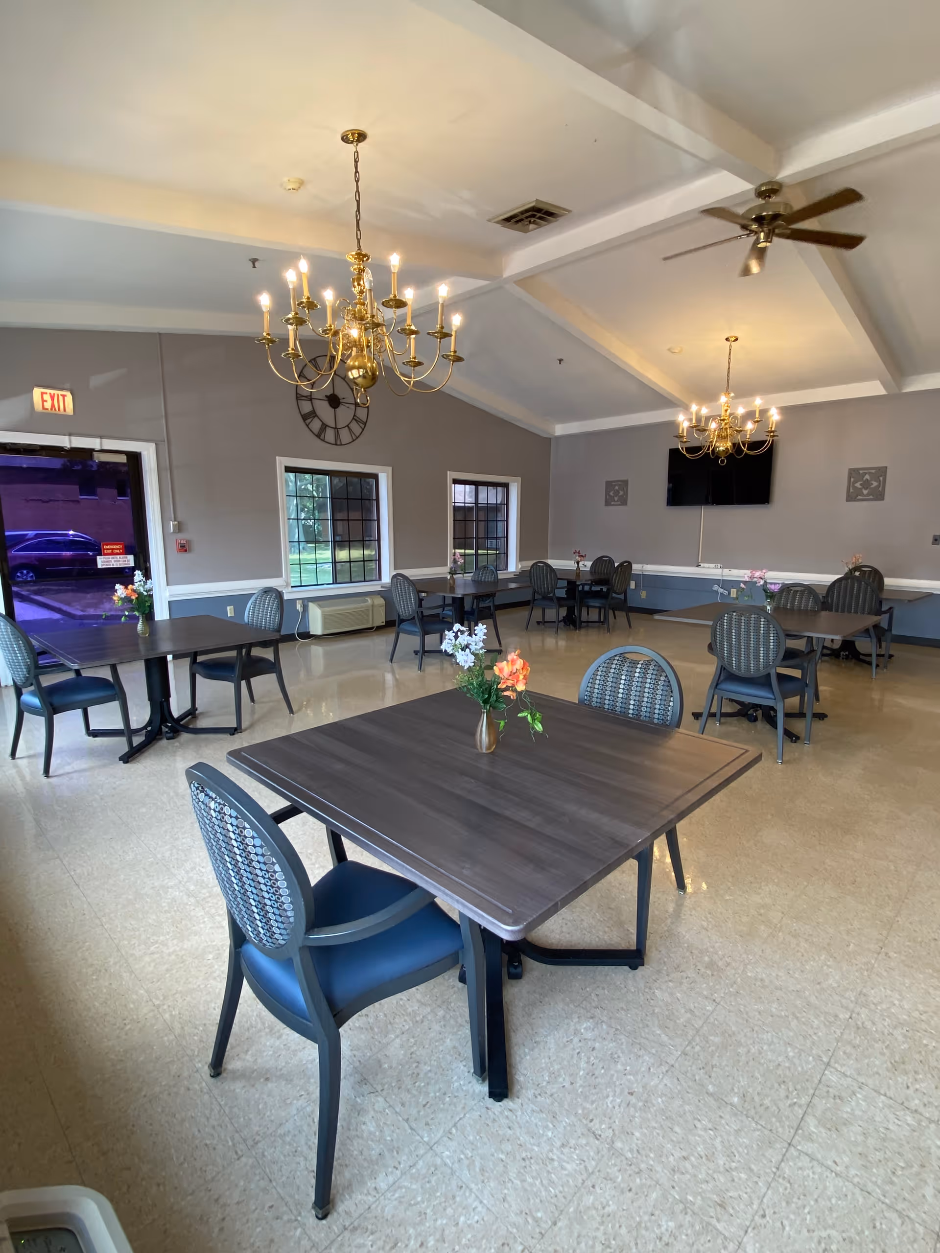 A dining room with several square tables and chairs arranged neatly. Each table has a small vase with flowers. The room features two ornate chandeliers hanging from a vaulted ceiling, a large wall clock, and a flat-screen TV mounted on the wall. There are windows letting in natural light and an exit door visible on the left side.