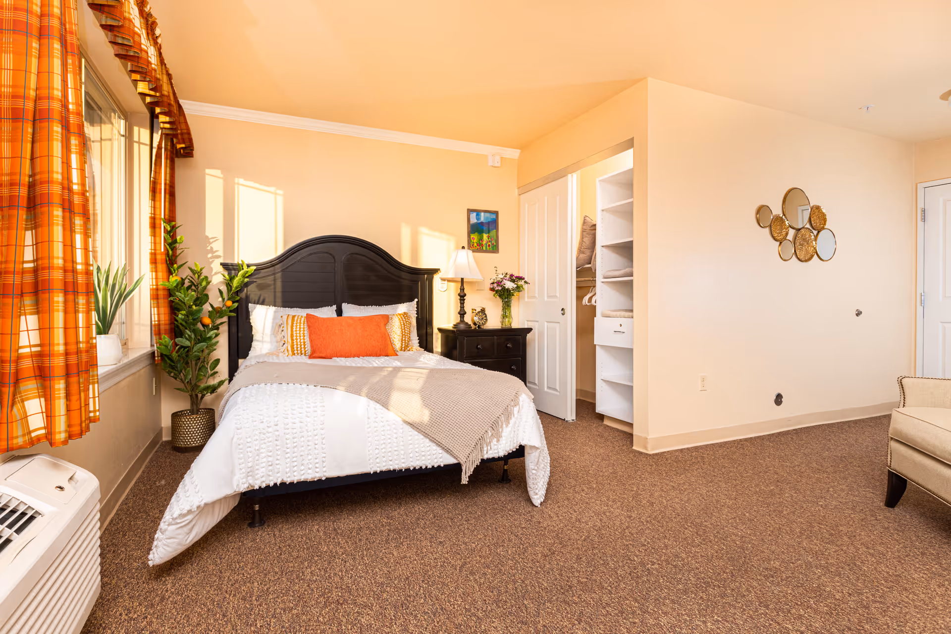 A bright and cozy bedroom with a black headboard bed dressed in white and beige bedding with orange accent pillows. There is a nightstand with a lamp and flowers, a closet with sliding doors and shelves, a beige armchair, and a window with orange plaid curtains letting in sunlight. The walls are light beige and the carpet is brown.