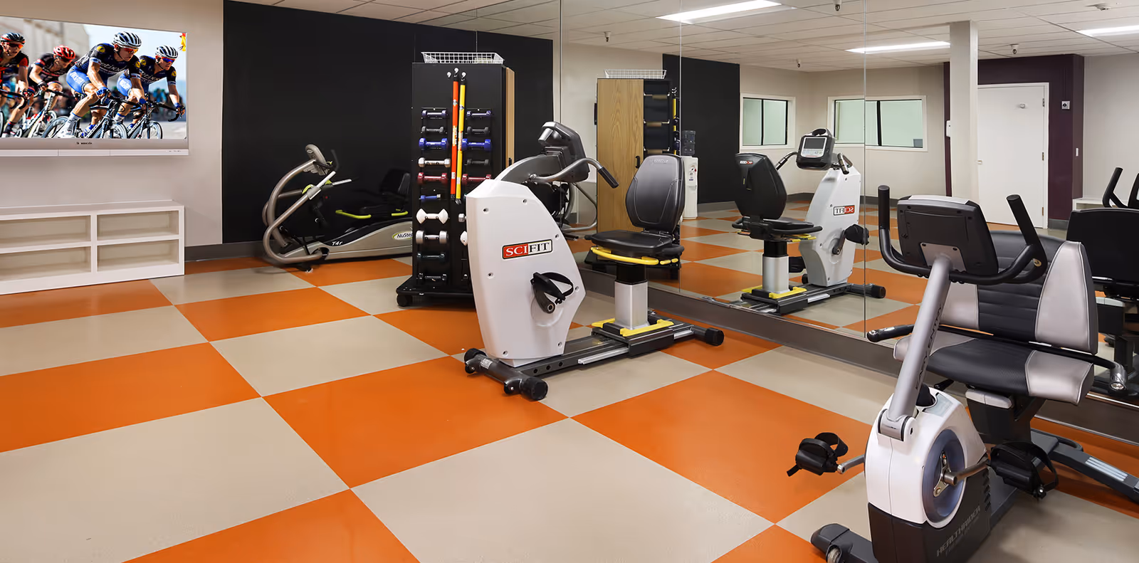 Exercise room with recumbent bikes, weight rack, mirrored wall and orange-and-beige checkered floor.