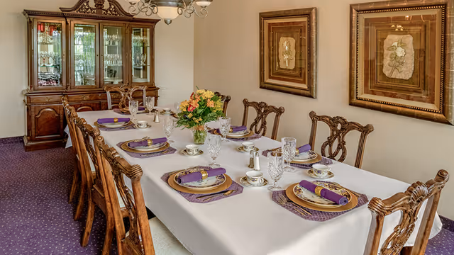 A formal dining room with a long table covered with a white tablecloth set for eight people. Each place setting includes a plate, a purple napkin with a gold napkin ring, a teacup, and a crystal glass. A flower arrangement is placed in the center of the table. The room has wooden chairs with intricate designs, a wooden china cabinet filled with glassware, and two framed floral artworks on the wall.