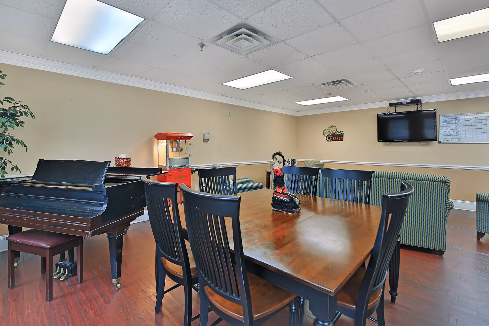 Communal activity room with a large wooden dining table and chairs, a grand piano, striped sofas, a wall-mounted TV and a popcorn machine.