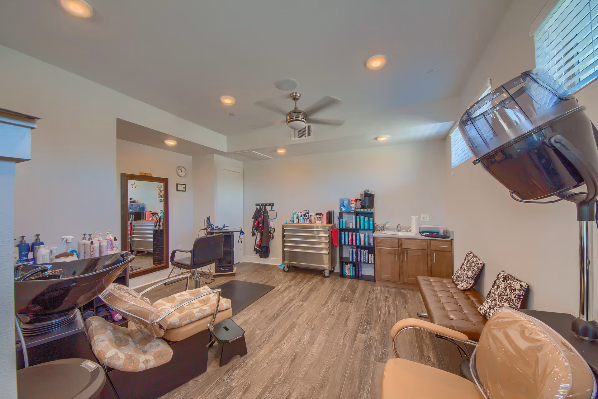 Interior of a hair salon room with a shampoo station, styling chair, large mirror, hair care products on shelves and a cabinet, a ceiling fan, and a brown cushioned bench with pillows. The room has wood flooring and white walls with windows near the ceiling.