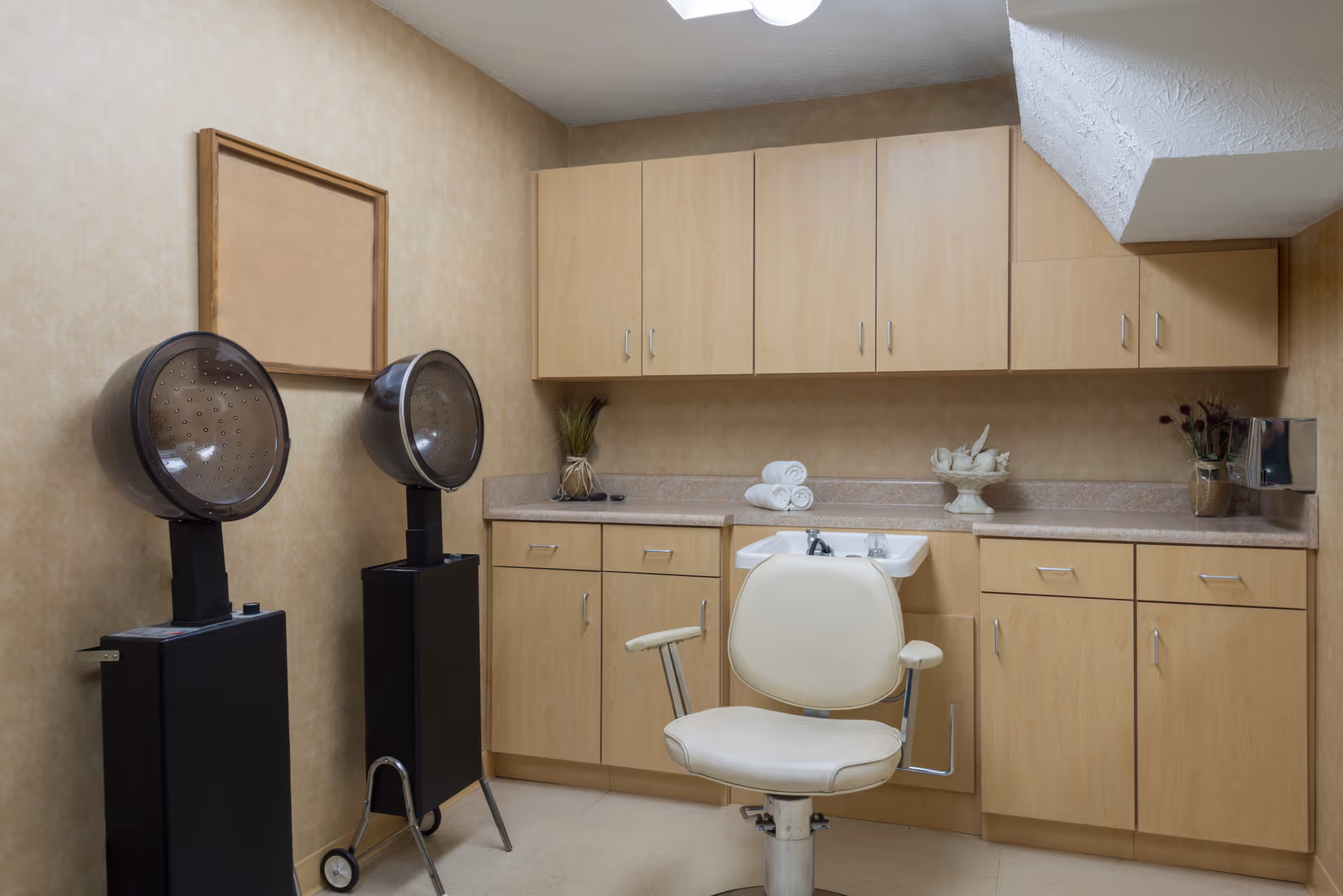 Interior view of a salon or beauty treatment room with two black hair dryers on stands, a white salon chair in front of a small sink, light wood cabinets above and below the countertop, and decorative items including rolled white towels, a vase with flowers, and a decorative bowl.