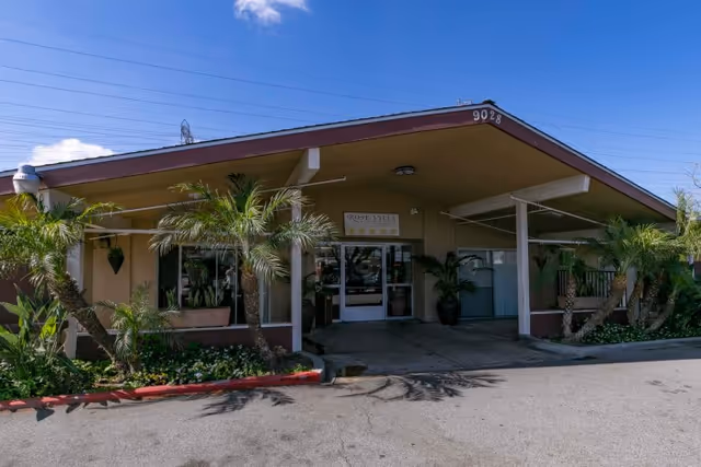 Front exterior view of Rose Villa Healthcare Center building with a slanted roof, palm trees, and plants along the entrance driveway under a clear blue sky.