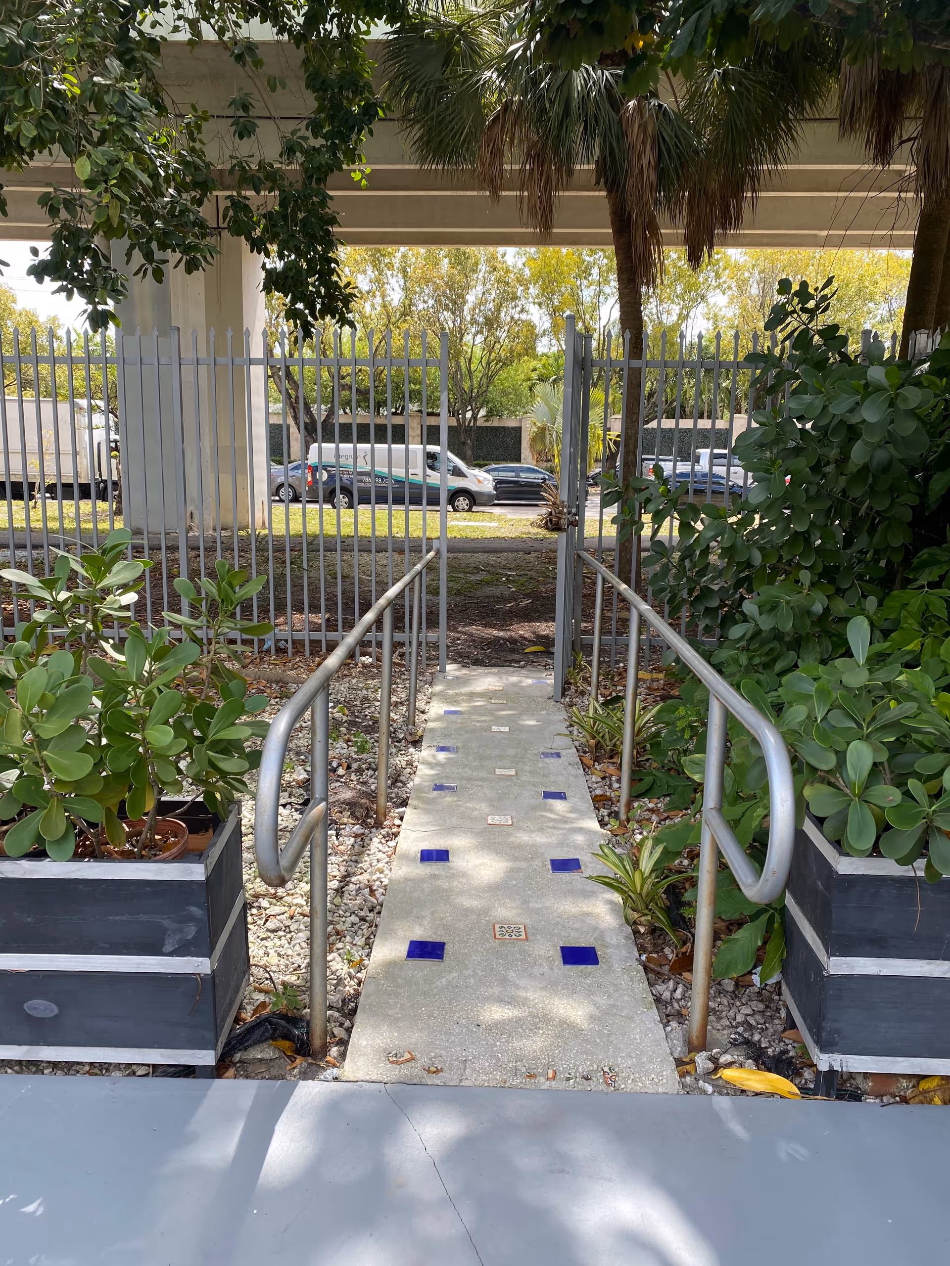 A narrow concrete pathway with blue and white tiles embedded in it, flanked by metal handrails on both sides. The path is bordered by green plants in large black planters and leads to a metal gate with a fence. Trees and vehicles are visible in the background under an overpass.