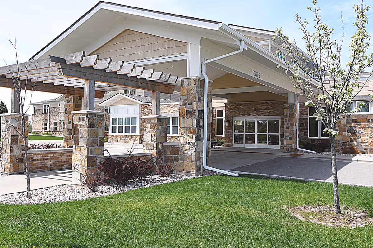 Entrance of a senior living facility with a covered drop-off area supported by stone pillars and a wooden pergola. The building exterior features stone and brickwork with multiple windows. There is a small tree and green grass in the foreground.