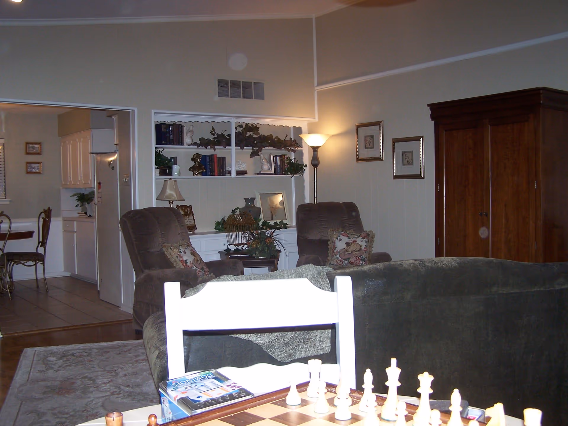A cozy living room with two brown armchairs, a dark green sofa, a wooden armoire, and a built-in white bookshelf decorated with books, plants, and ornaments. A floor lamp and two framed pictures hang on the wall. In the foreground, a chessboard and a dominoes game box are visible on a table. The living room opens into a kitchen area with white cabinets and a dining table with chairs.