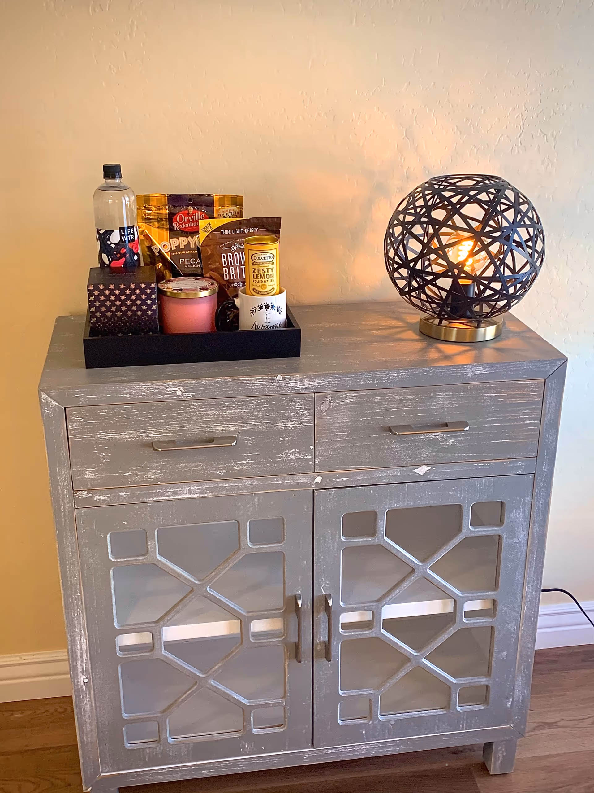 A rustic gray wooden cabinet with two drawers and two glass-paneled doors. On top of the cabinet, there is a black tray holding a bottle of water, snack bags, a candle, and a small container. To the right of the tray, there is a decorative spherical lamp with a geometric metal frame emitting a warm light. The cabinet is placed against a beige wall and on a wooden floor.