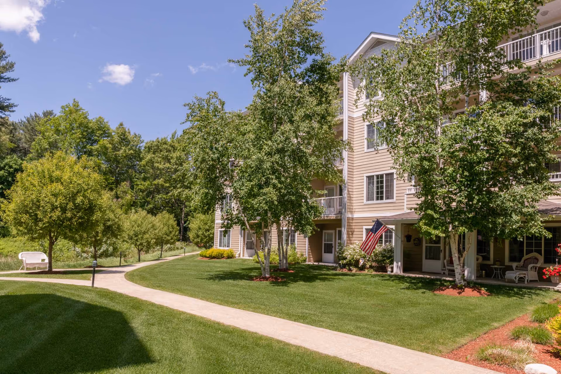 Exterior view of a multi-story residential building with beige siding, white trim, and balconies. The building is surrounded by green grass, trees, and a paved walkway. An American flag is displayed near the entrance, and there is outdoor seating on a covered porch. The sky is clear with a few clouds.