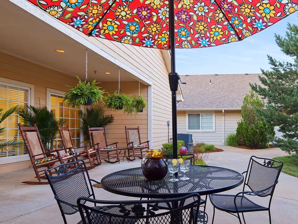 Outdoor patio area at Park West Plaza featuring a round black metal table with a colorful floral umbrella, a pitcher of iced tea with lemon slices, and three glasses. Surrounding the patio are several wooden rocking chairs with cushions, hanging green plants, and beige building walls with windows. Trees and shrubs are visible in the background under a clear sky.