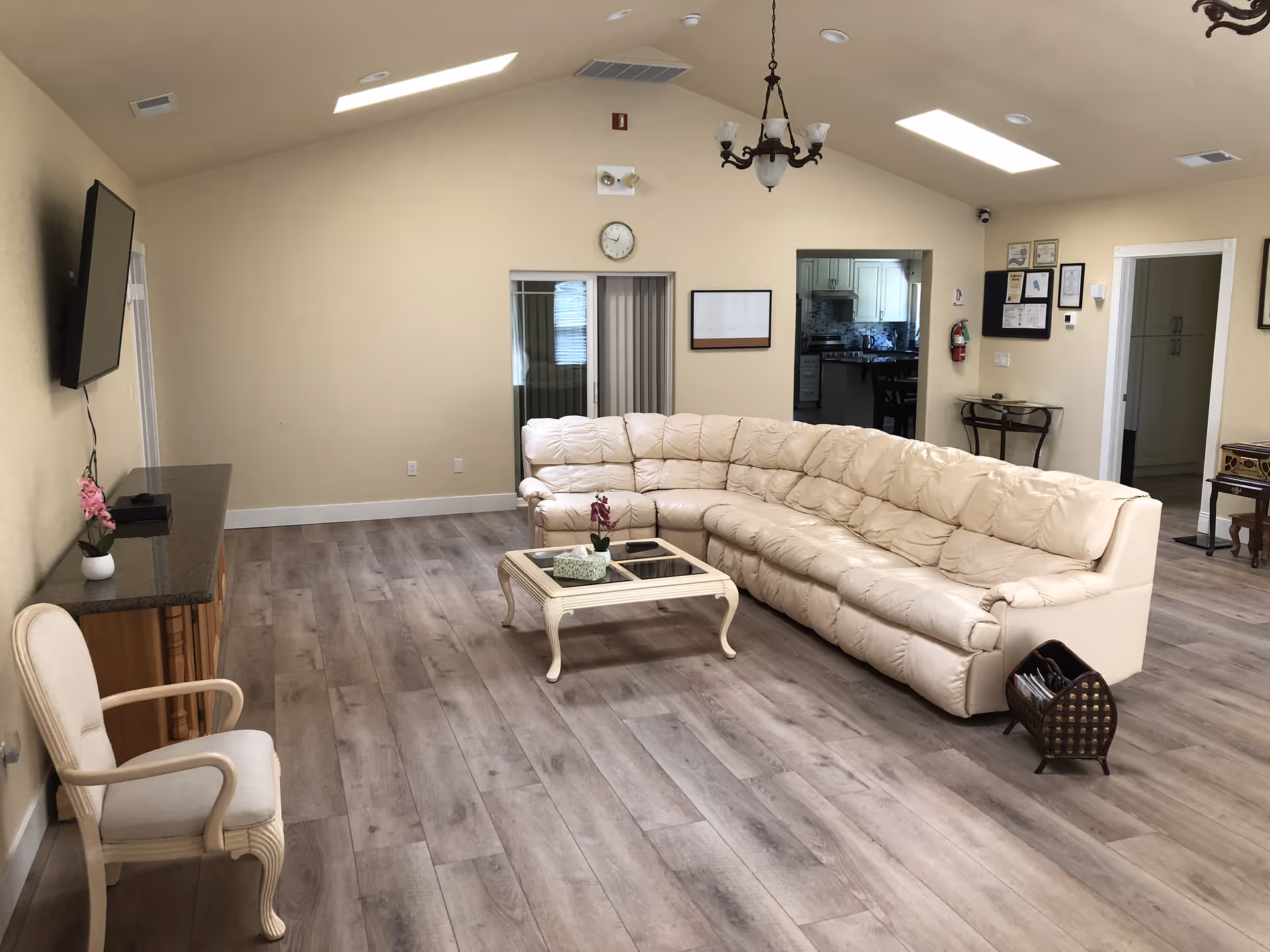 A spacious living room with a large cream-colored sectional sofa arranged around a glass-top coffee table. The room has light wood flooring, beige walls, a wall-mounted flat-screen TV, a single armchair, and a chandelier hanging from the ceiling. There is a clock on the far wall above a sliding door, and an open doorway leading to a kitchen area.