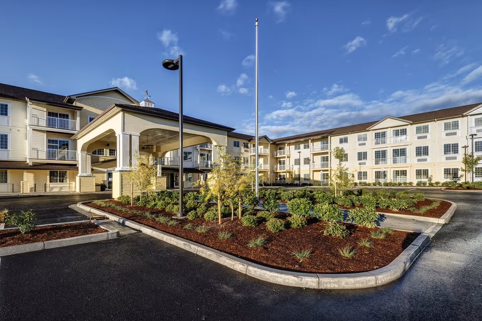 Front exterior of a three-story senior living building with a covered entrance, circular driveway and landscaped island under a blue sky.