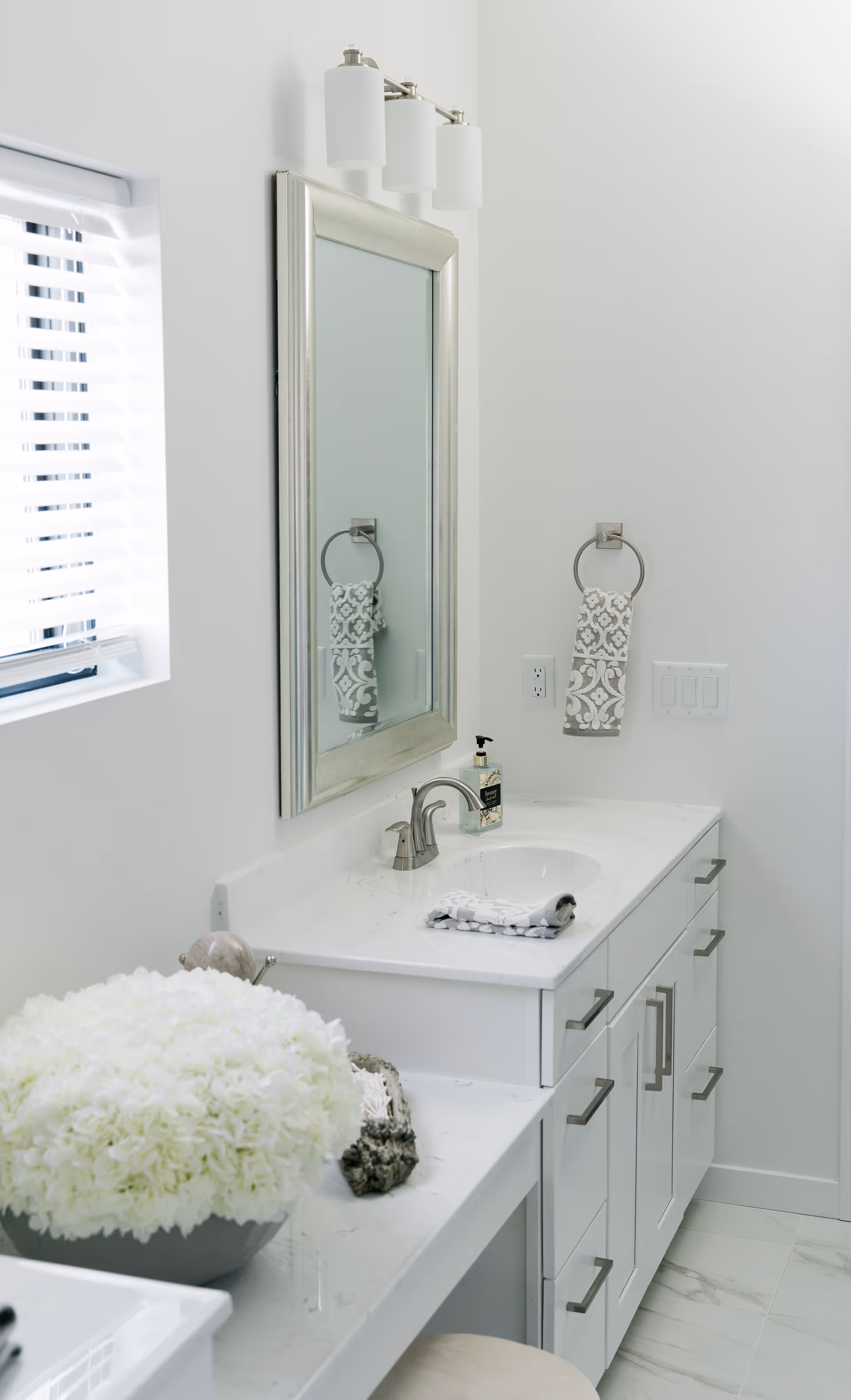 Bright and clean bathroom vanity area with a white countertop, a silver-framed mirror, a modern faucet, a patterned hand towel on a ring holder, a soap dispenser, and a large bowl of white flowers on the counter.