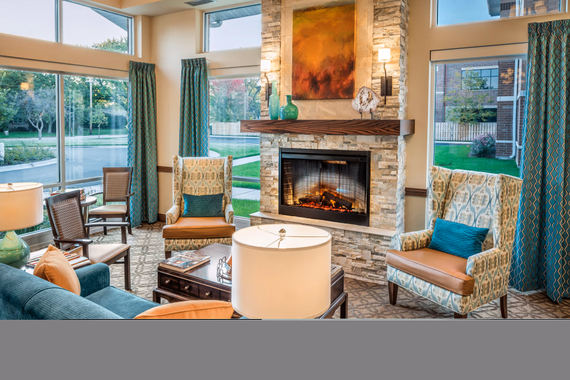 A cozy living room area with a stone fireplace featuring a lit fire. The room has large windows with teal patterned curtains, two patterned armchairs with teal cushions, a teal sofa with orange pillows, a wooden coffee table, and a white table lamp. Outside the windows, greenery and a building are visible.