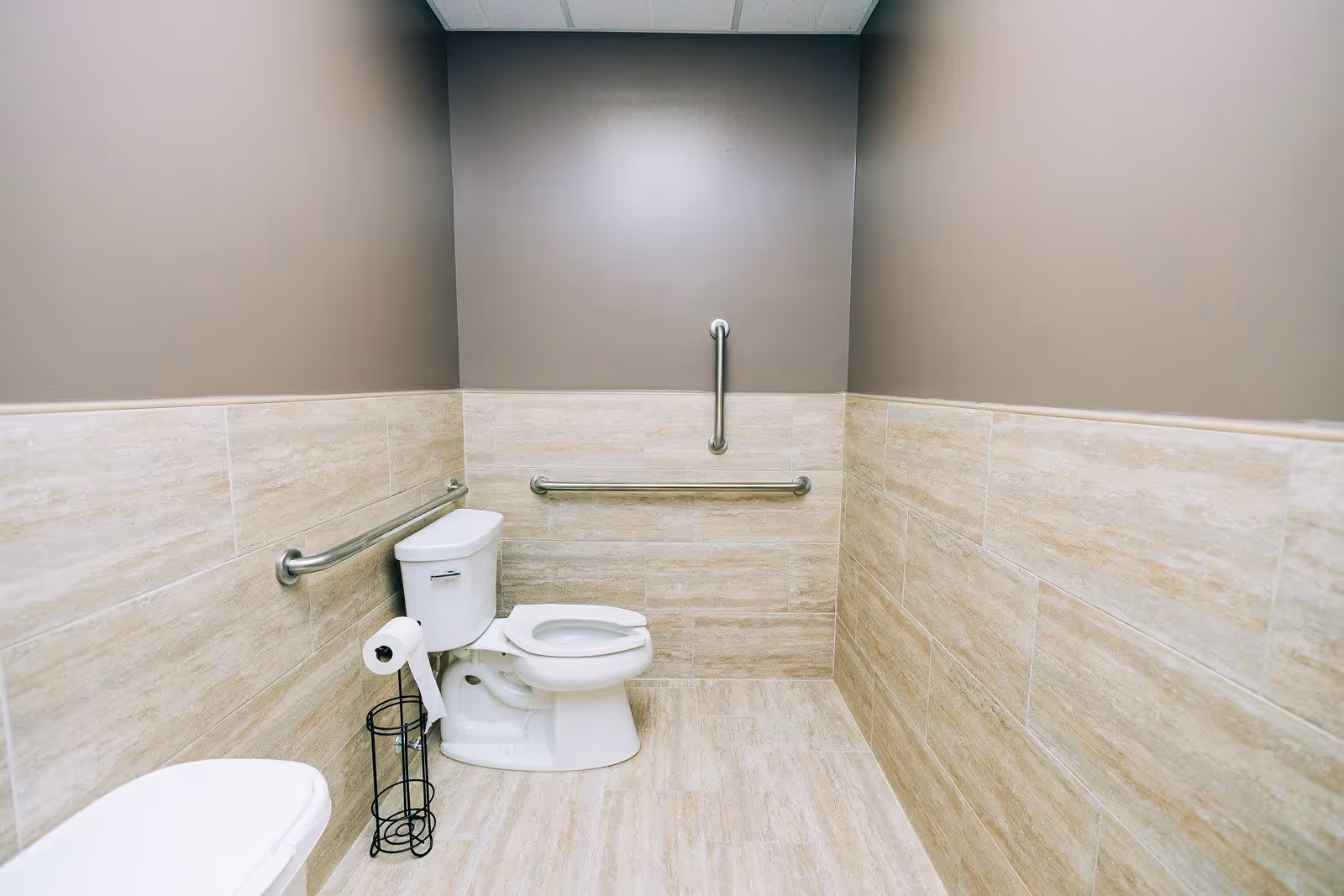 A clean, accessible bathroom with beige tiled walls and floor, featuring a white toilet with grab bars on the walls for support and a toilet paper holder next to it.