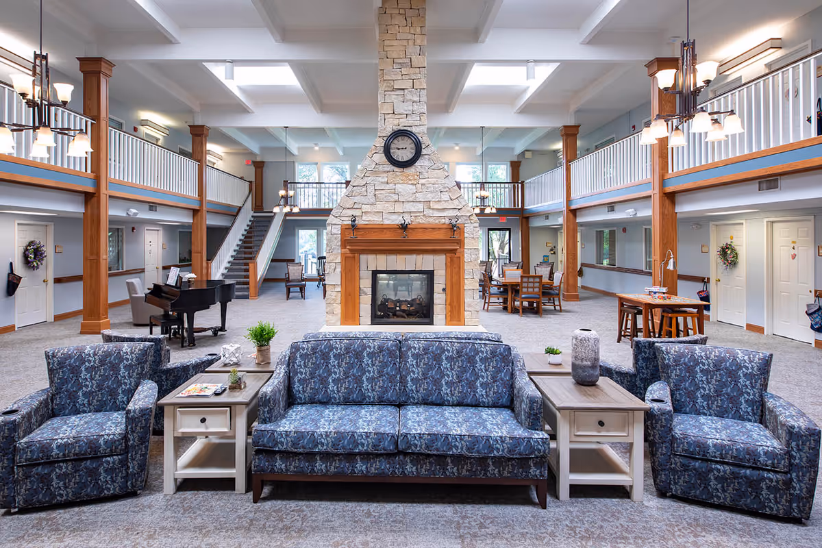 Spacious and well-lit assisted living facility common area with a large stone fireplace in the center, a clock mounted above it, and comfortable blue patterned seating including a sofa and armchairs. There are wooden columns, a grand piano to the left, dining tables and chairs in the background, and a staircase leading to an upper balcony with white railings. The room has carpeted floors and multiple light fixtures hanging from the ceiling.