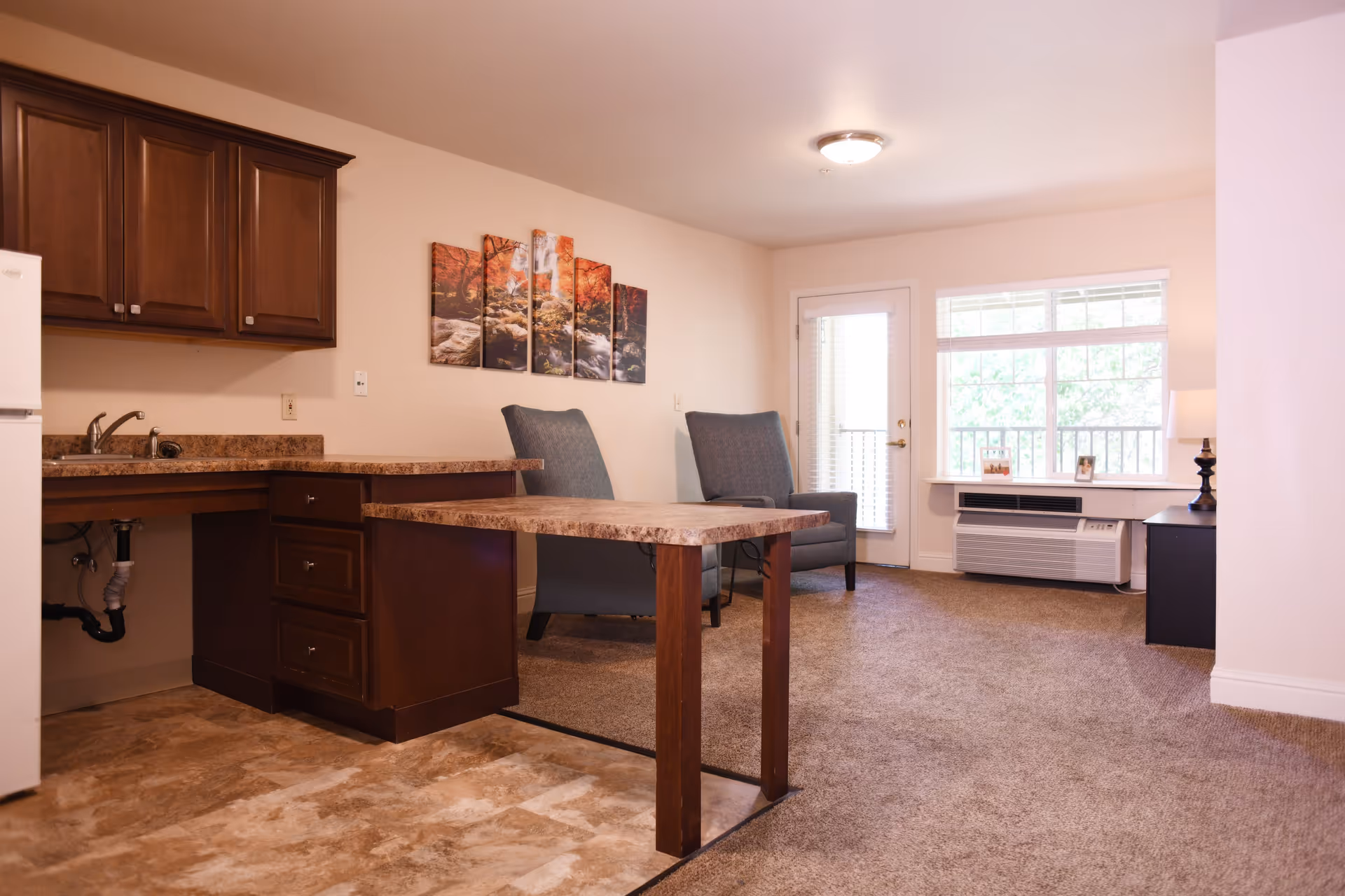 Interior view of a living space in Oakdale Heights Assisted Living featuring a small kitchen area with wooden cabinets and a countertop, two upholstered armchairs, a window with blinds, a door leading outside, and a small table with a lamp and framed photos.