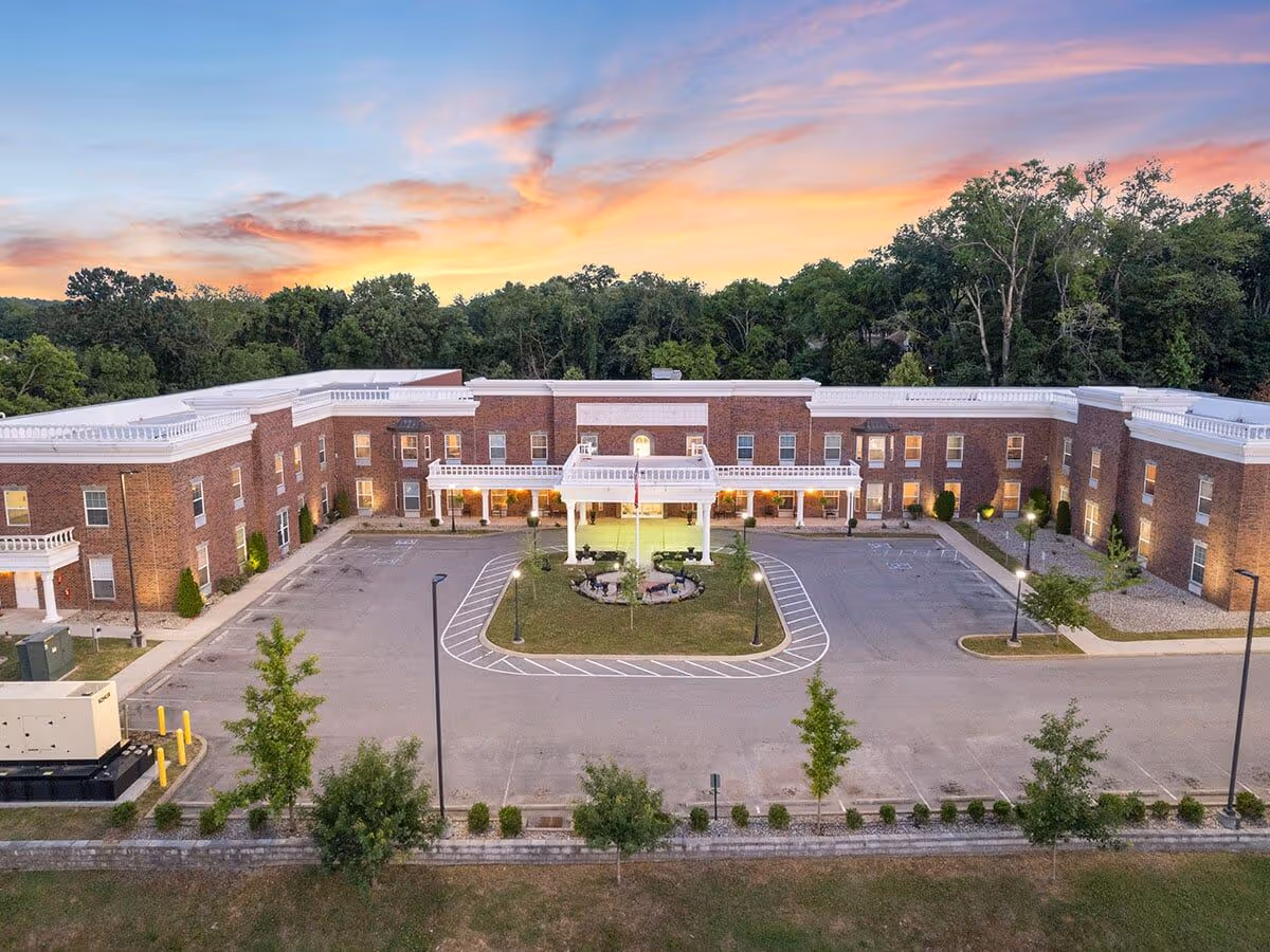 Aerial view of a two-story brick senior living building with a circular driveway, landscaped center island, and trees at sunset.