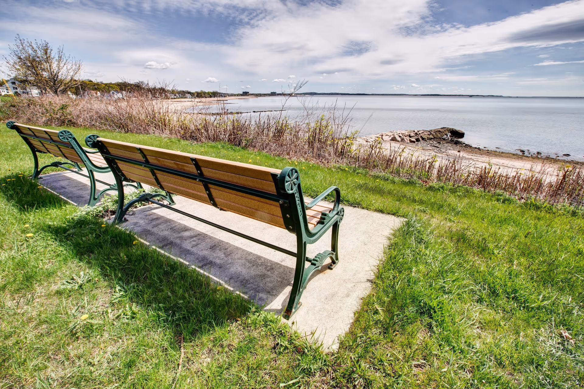 Two empty wooden benches with green metal frames sit on a concrete slab overlooking a calm body of water with a rocky shoreline and grassy area under a partly cloudy sky.