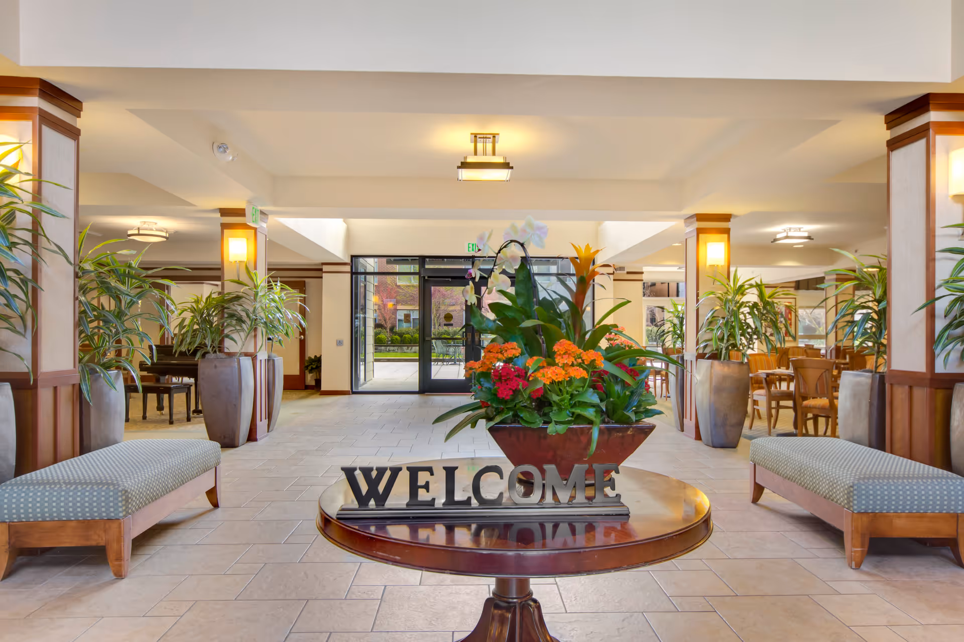 Bright and spacious lobby area with a round wooden table in the center holding a flower arrangement and a decorative 'WELCOME' sign. The lobby features tiled flooring, cushioned benches on either side, large potted plants, and glass doors leading outside. Warm lighting and wooden accents create a welcoming atmosphere.