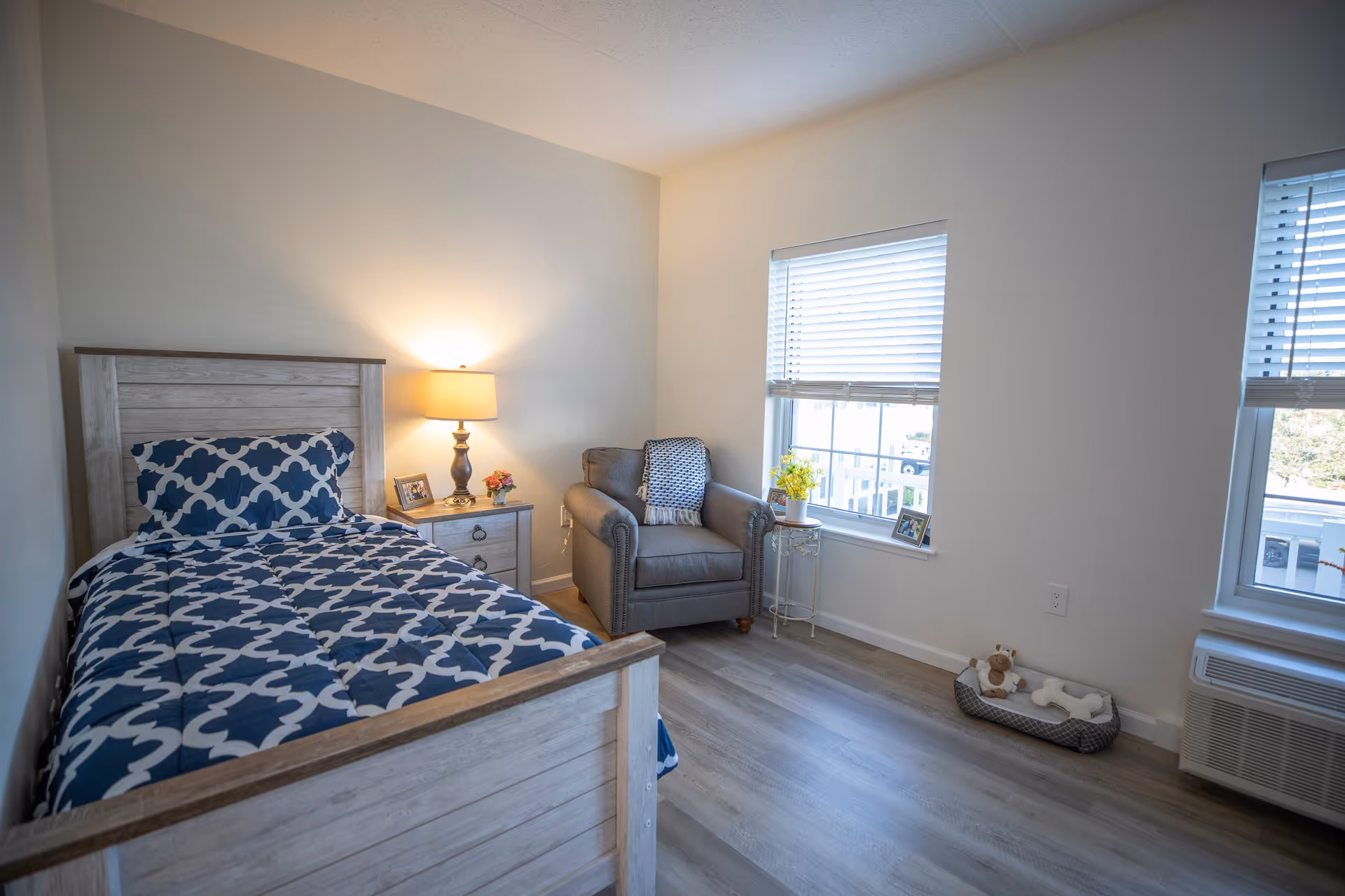 A bright and cozy bedroom with a single bed featuring a blue and white patterned comforter, a wooden headboard and footboard, a nightstand with a lamp, framed photos, and a small flower vase. Next to the nightstand is a comfortable gray armchair with a blanket draped over it. Two windows with white blinds let in natural light, and a small pet bed with toys is placed near the wall. The room has light-colored walls and wood flooring.
