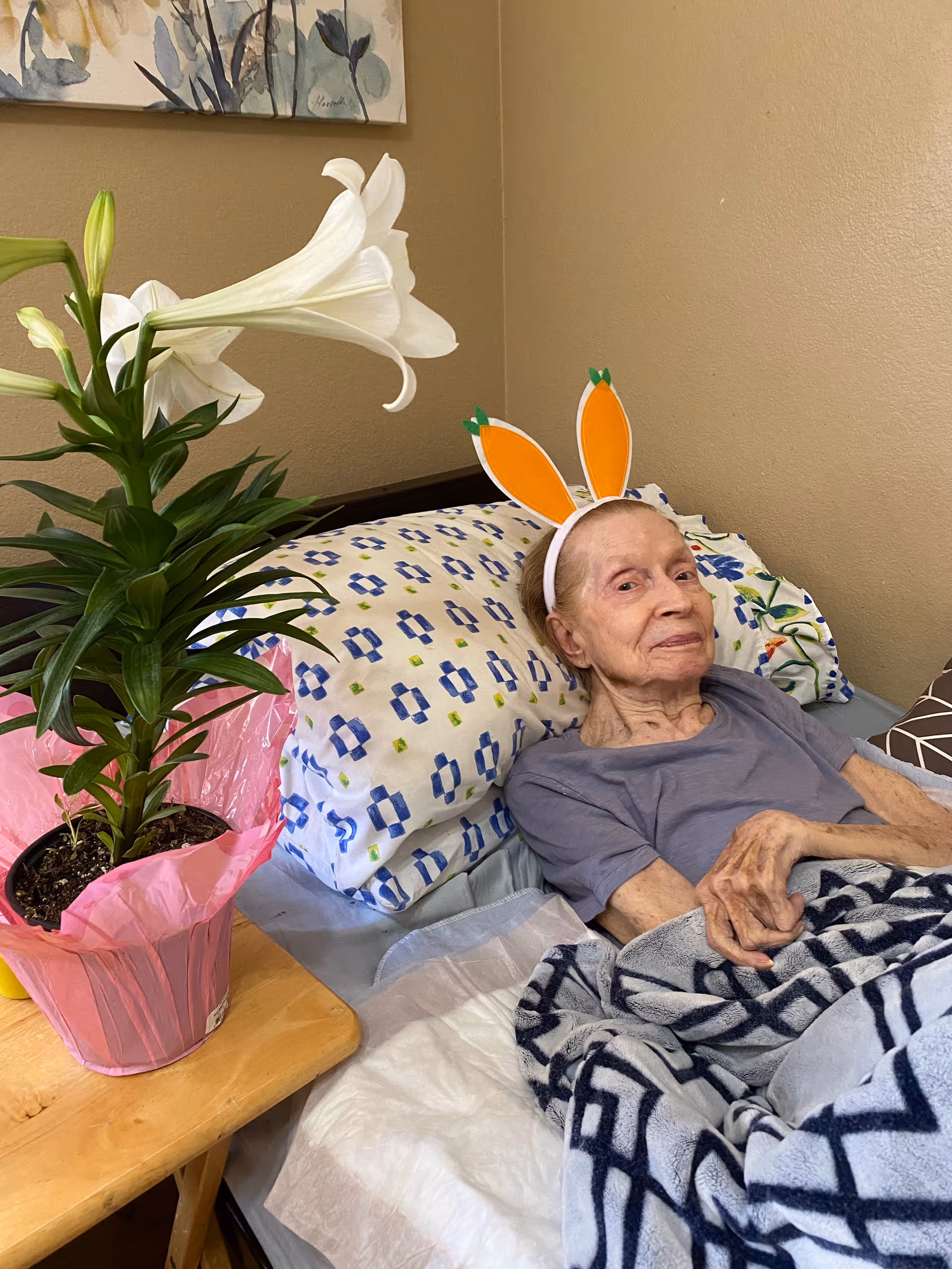 An elderly person lying in bed wearing a headband with orange bunny ears. There is a potted white lily plant wrapped in pink paper on a small wooden table next to the bed. The bed has patterned pillows and a blanket with geometric designs. The room has beige walls and a floral painting is partially visible above the bed.