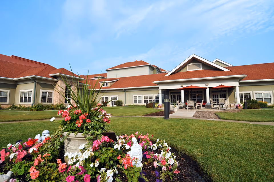 Exterior view of The Oaks at Bartlett senior living facility with a well-maintained lawn, colorful flower bed in the foreground, and a building with red roofs and a covered patio area with tables and chairs under red umbrellas.
