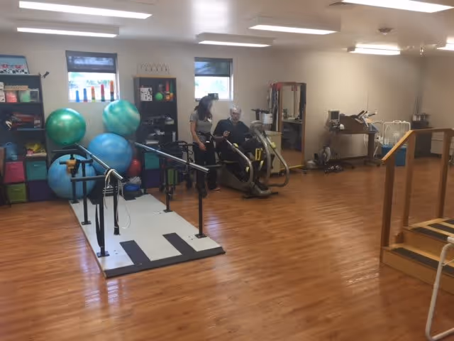 A spacious rehabilitation or physical therapy room with wooden floors, exercise equipment including parallel bars, large exercise balls, and a seated exercise machine. A caregiver is assisting an elderly man using the exercise machine. Shelves with colorful bins and therapy tools are visible against the wall.