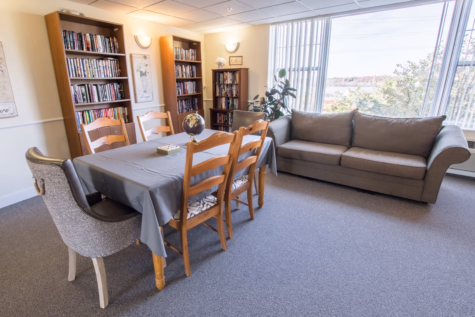 A cozy common room with a gray sofa near large windows with vertical blinds, a table covered with a gray tablecloth surrounded by wooden chairs, and bookshelves filled with books against the wall.