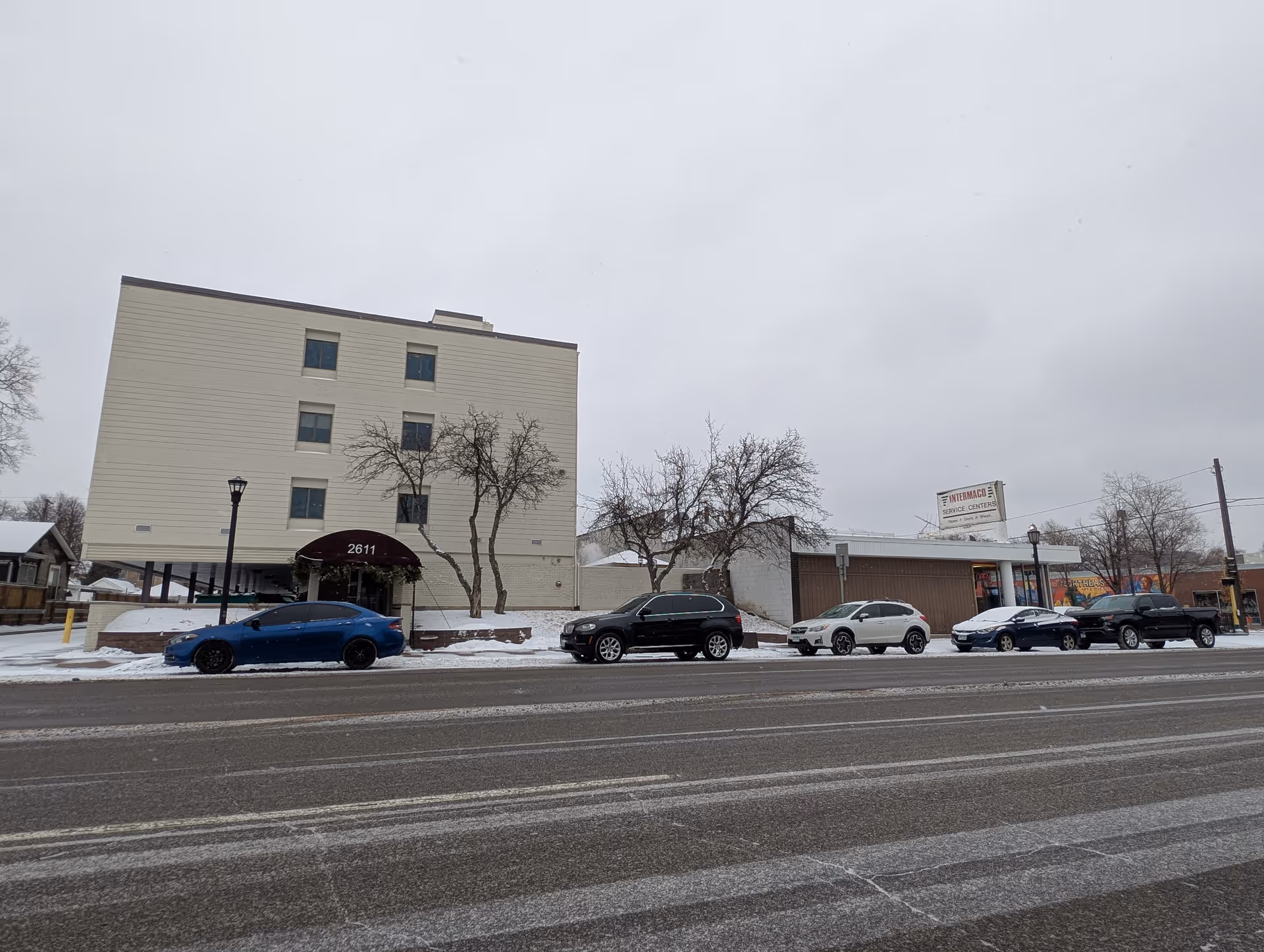 Four-story beige building with a maroon awning reading '2611' and several cars parked along a snowy street in front.