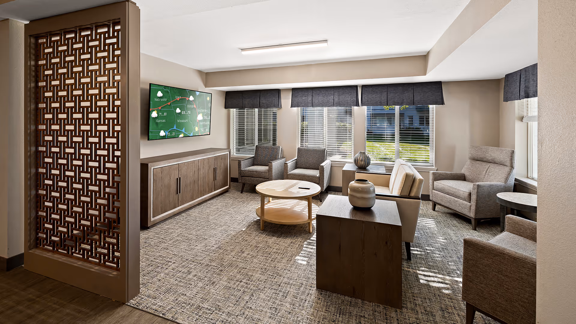 A bright and modern living room area with multiple armchairs and a sofa arranged around a round wooden coffee table and a wooden side table with decorative vases. Large windows with blinds and dark valances let in natural light. A flat-screen TV is mounted on the wall above a wooden cabinet. A decorative wooden partition is visible on the left side of the room.